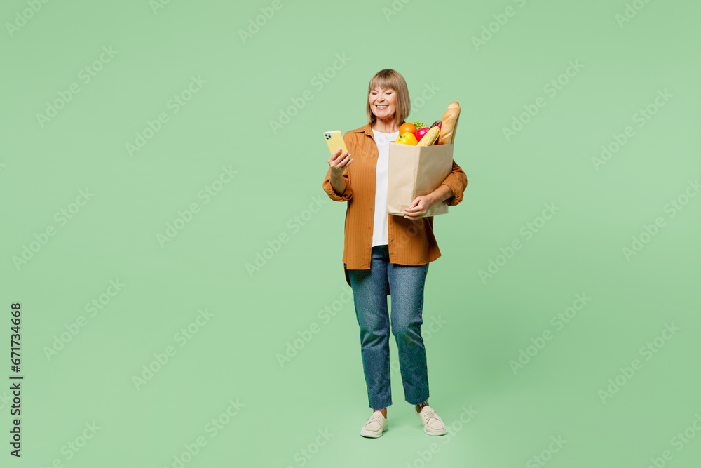 Full body elderly woman wear brown shirt casual clothes hold shopping paper bag with food product use mobile cell phone browsing internet isolated on plain green background Delivery service from shop Full body elderly woman wear brown shirt casual clothes hold shopping paper bag with food product use mobile cell phone browsing internet isolated on plain green background Delivery service from shop