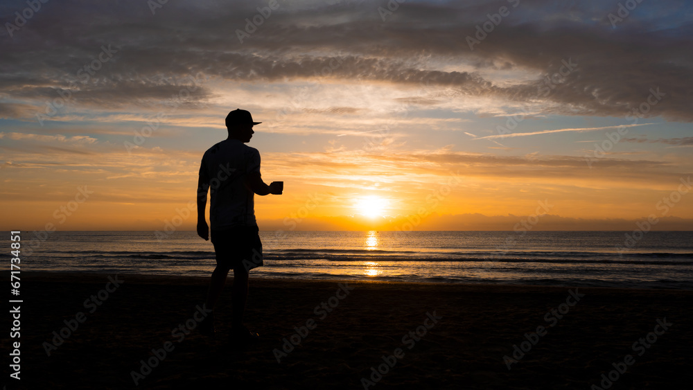 Man standing on the beach against light, with cup of coffee in his hand and cap. sunrise, beach, figure against light Man standing on the beach against light, with cup of coffee in his hand and cap. sunrise, beach, figure against light