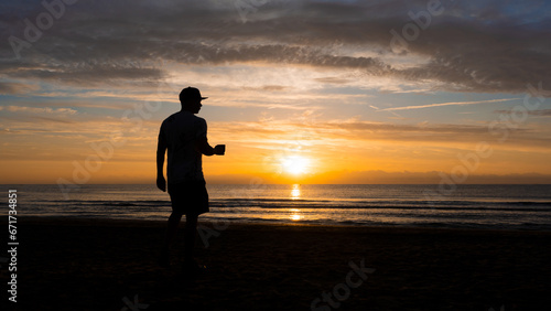 Man standing on the beach against light, with cup of coffee in his hand and cap. sunrise, beach, figure against light