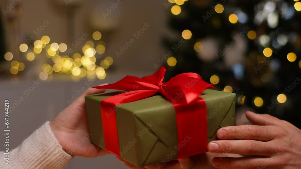 Close-up cropped shot of unrecognizable happy young woman receiving Christmas gift from loving man at home on blurred background of decorating xmas tree, celebration bokeh lights, slow motion. Close-up cropped shot of unrecognizable happy young woman receiving Christmas gift from loving man at home on blurred background of decorating xmas tree, celebration bokeh lights, slow motion.