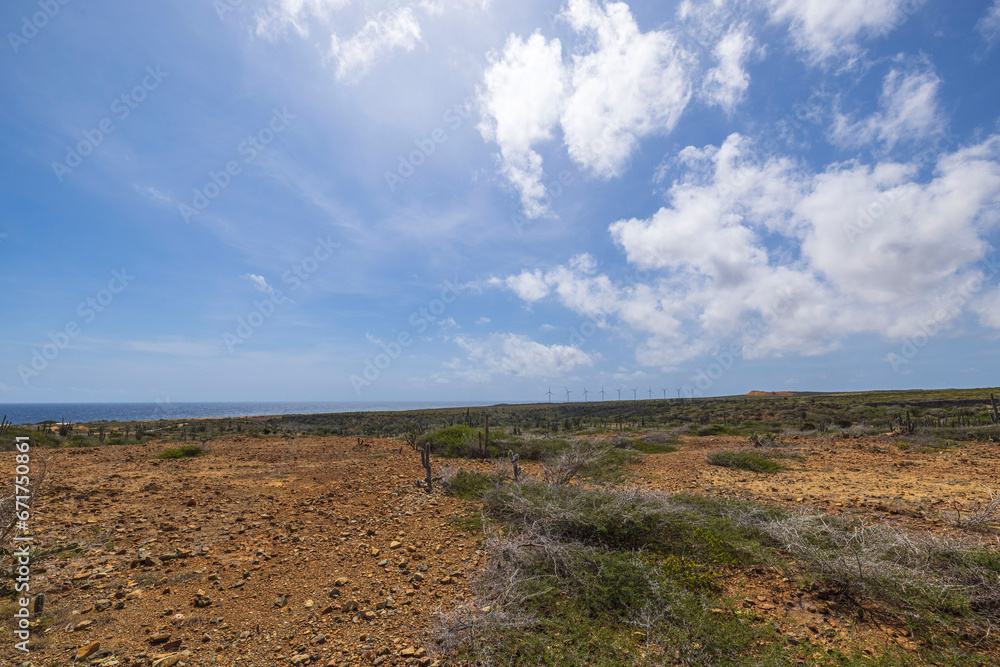 Fototapeta premium Stunning view of rocky desert set against backdrop of Caribbean sea and clear blue sky with white clouds with wind turbines visible in distance. Aruba.