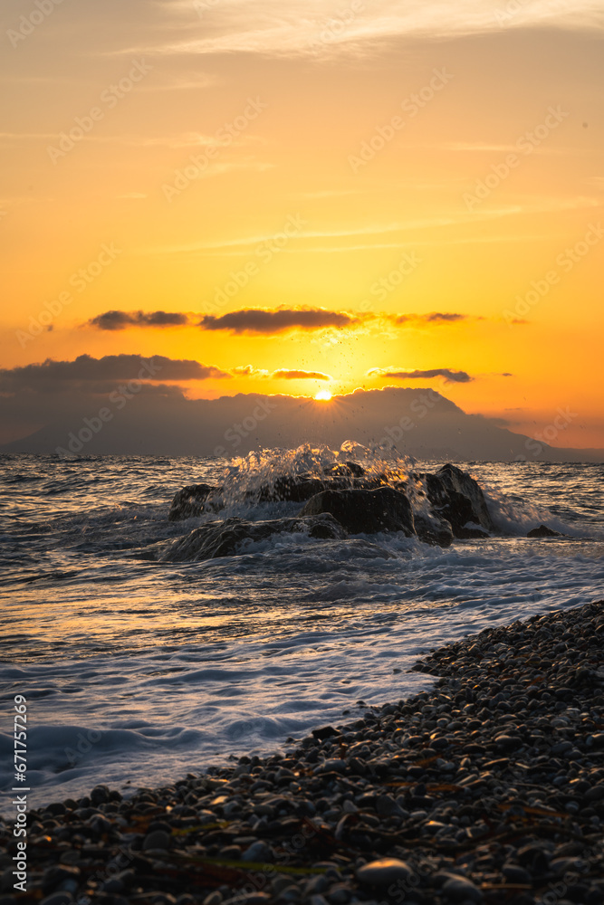 Sunrise over the ocean. The sun is a golden orange color partially obscured by clouds. In the foreground are rocky beaches with large boulders and in the background are the Greek mountains.