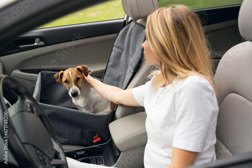 Road trip by car. Petting cute dog friend while stop in car trip. Dog sitting in car chair hammock on front seat of the car. Blonde young driver girl petting her pet. Focus on dog, girl is blurred