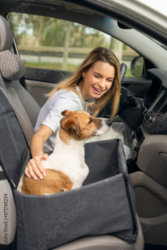 Happy smiling blonde girl in car. dog sitting front seat in car hammock. travel with your pet safely and comfortably. petting Jack Russell terrier and smiling. focus on girl, dog blurred out of focus