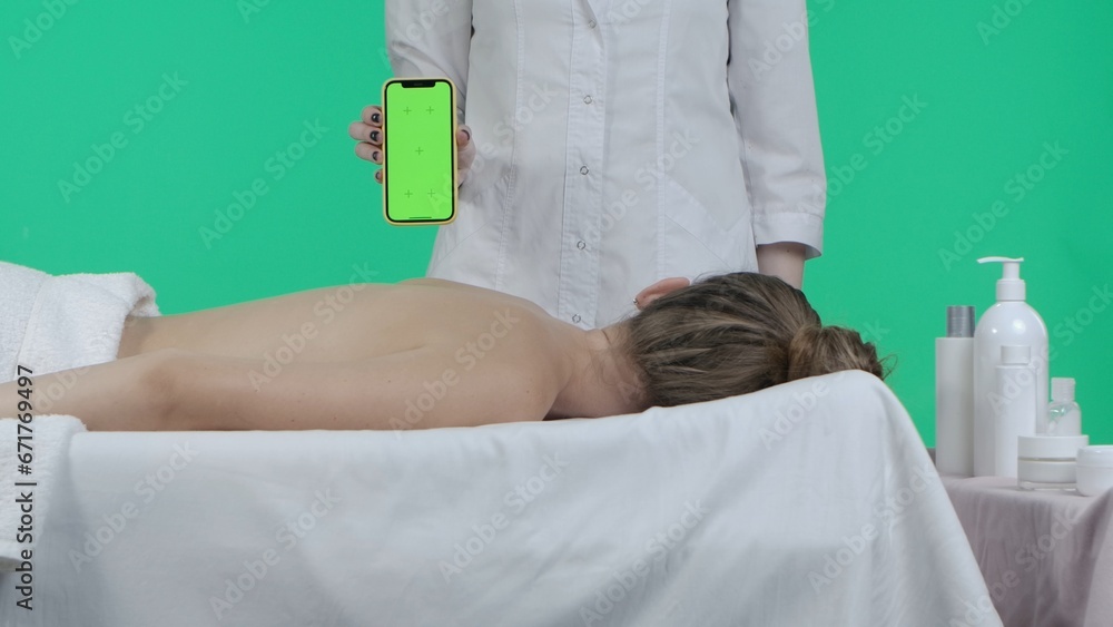 Obraz premium Beauty salon room, female laying on massage table, masseur standing near holding smartphone with Chroma key green screen background, advertising area workspace mockup.