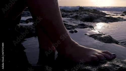 Traveling of bare feet of a woman sitting on some rocks in the sea at sunset