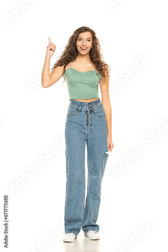 Full length shot of young smiling woman wearing green shirt and blue jeansi while standing at isolated white background and pointing up.