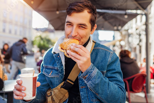 Young happy man having breakfast with coffee and croissant sitting at outdoor cafe in European city.