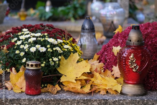 Grave with glass candles and flowers in pots among dry autumn leaves in cemetery on November day