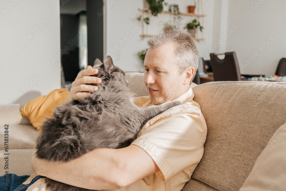 A middle-aged man sits with a gray cat on the sofa in the living room and relaxes after work. Adored pets A middle-aged man sits with a gray cat on the sofa in the living room and relaxes after work. Adored pets