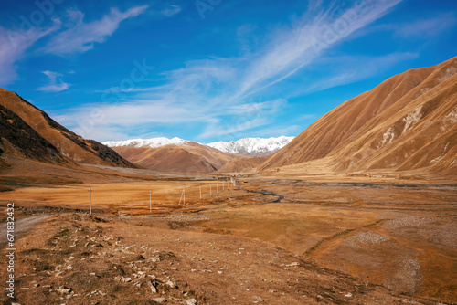 A vast mountainous landscape under a clear blue sky. Snow-capped peaks contrast with golden valley floors