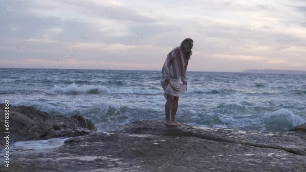 A woman walks on the rocks by the sea, her silk skirt is rolled by a wave A woman walks on the rocks by the sea, her silk skirt is rolled by a wave