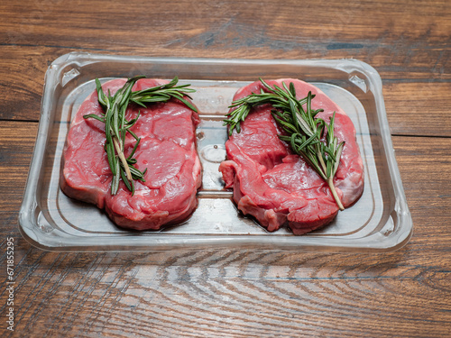 Juicy uncooked lamb leg steak with green fresh rosemary on plastic tray on wooden table. Uncooked high quality meat product with rich taste and vivid color.