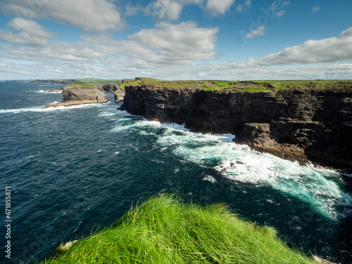 Edge of a cliff and powerful ocean hits rough stone coastline. low cloudy sky, warm sunny day. Kilkee area, Ireland. Travel, tourism and sightseeing concept. Irish landscape and coastline.