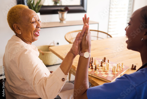 Black people, nurse and high five in elderly care for chess, fun or social activity or game together at home. African medical professional touching hands with senior female person with a disability photo