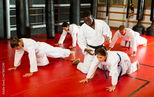 Man and women in kimono doing push-ups in gym during karate training. African-american man trainer correcting their moves.
