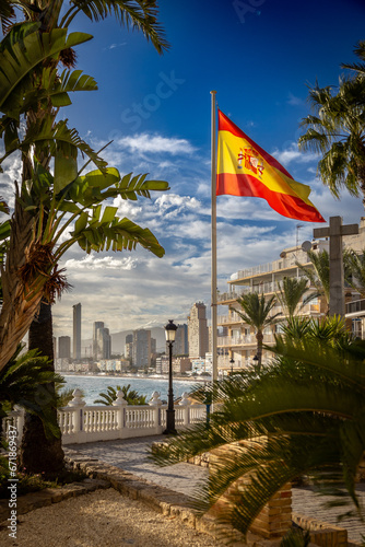 Flaga Hiszpanii na placu między palmami, a w tle widok na Hiszpańskie miasto Benidorm na Costa Blanca © Jakub