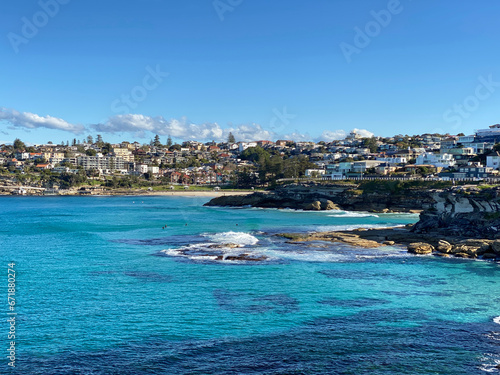Fototapeta Naklejka Na Ścianę i Meble -  Bay and coastline next to the ocean. View of the city in the distance. View of turquoise waves breaking on the shore. Rocky coast of the ocean. Landscape and shore. Australia, Sydney.