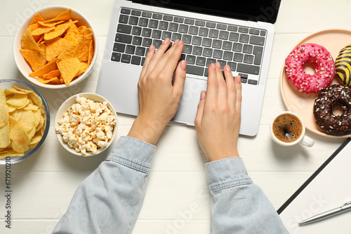 Bad eating habits. Woman using laptop surrounded by different snacks at white wooden table, top view