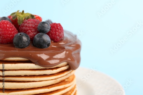 Stack of tasty pancakes with fresh berries and chocolate spread on light blue background, closeup. Space for text
