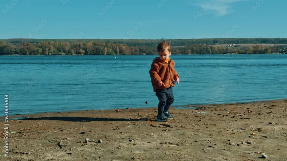 Male toddler wearing brown jacket spending time near the river on sunny day. Kid picks up a stone and runs to throw it into water. Male toddler wearing brown jacket spending time near the river on sunny day. Kid picks up a stone and runs to throw it into water.