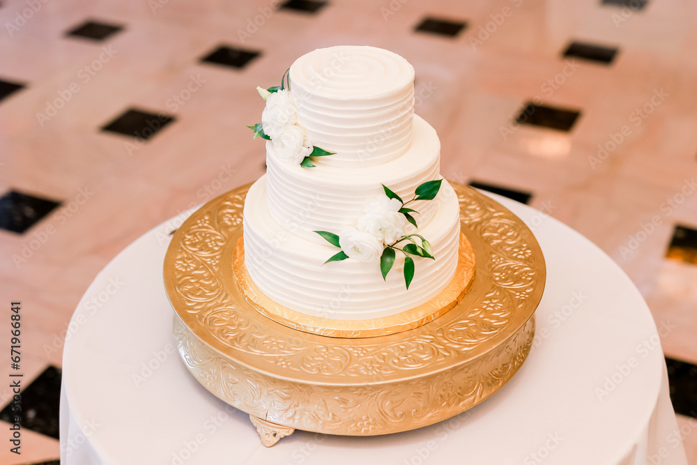 A white wedding cake with flowers and greenery on a gold platter on a white tablecloth table A white wedding cake with flowers and greenery on a gold platter on a white tablecloth table