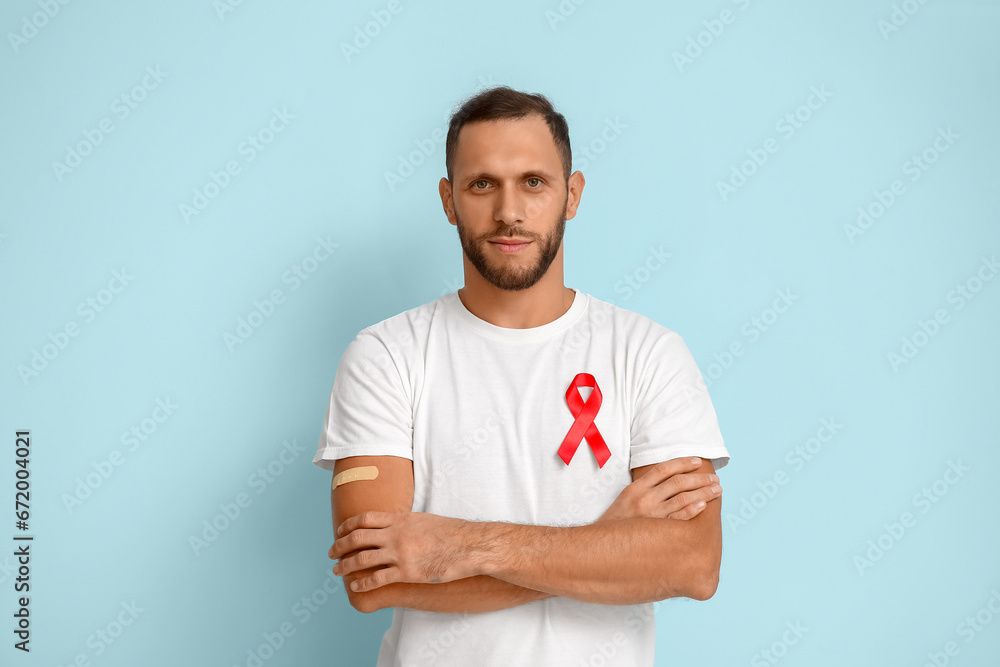 Young man with red ribbon and crossed arms on blue background. AID awareness concept - obrazy, fototapety, plakaty 