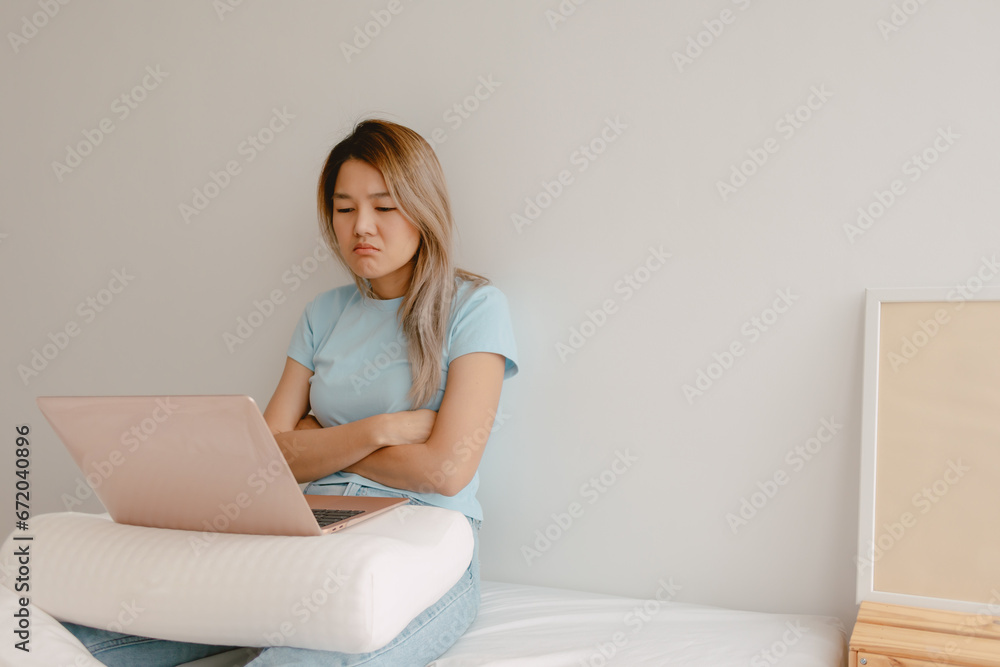 Unhappy asian Thai woman dislike and bored face, using laptop for working at home, putting computer on pillow lap, sitting on white bed, worried trouble at apartment room alone. Unhappy asian Thai woman dislike and bored face, using laptop for working at home, putting computer on pillow lap, sitting on white bed, worried trouble at apartment room alone.