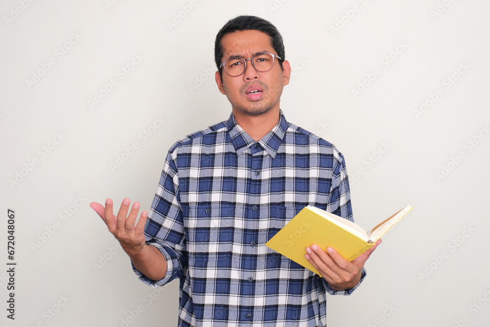 Adult Asian man holding a book with confused expression Adult Asian man holding a book with confused expression