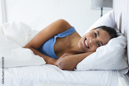 Portrait of happy biracial woman wearing blue pyjamas and lying in bedroom photo