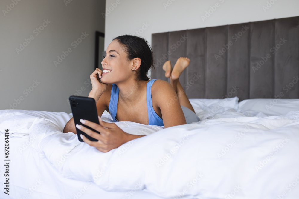 Happy biracial woman using smartphone and lying in bedroom Happy biracial woman using smartphone and lying in bedroom