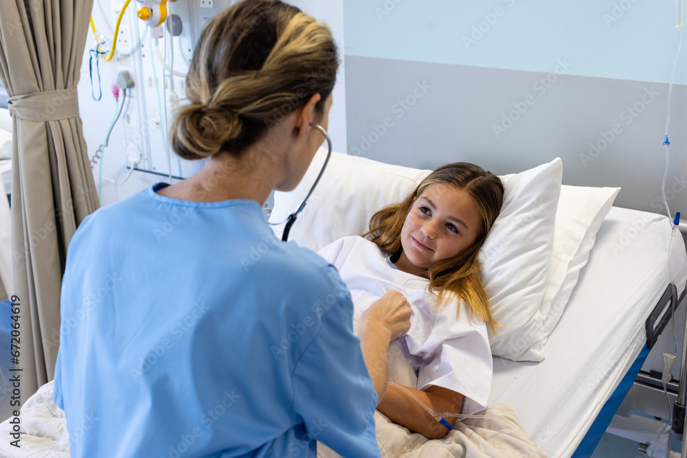Caucasian female doctor using stethoscope on chest of smiling girl patient lying in hospital bed Caucasian female doctor using stethoscope on chest of smiling girl patient lying in hospital bed