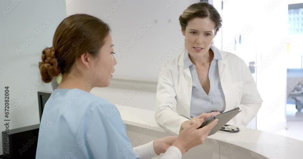 Focused diverse female doctors using tablet and discussing over hospital reception desk, slow motion Focused diverse female doctors using tablet and discussing over hospital reception desk, slow motion