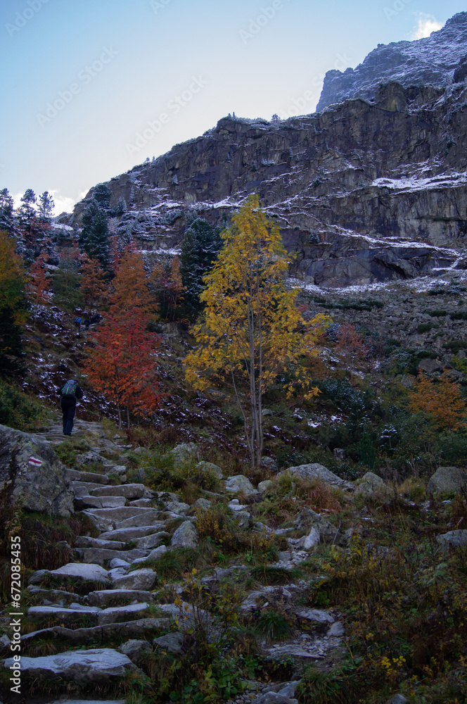 In Poland's High Tatras, a lone hiker treads on an ancient stone path, framed by vivid autumn leaves and a sprinkle of snow. Majestic cliffs complete this captivating scene.