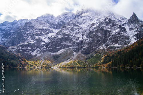 Scenic capture of Morskie Oko lake during fall. Snowy peaks tower behind, reflecting in clear waters. Vibrant autumn trees surround, creating a serene nature landscape.