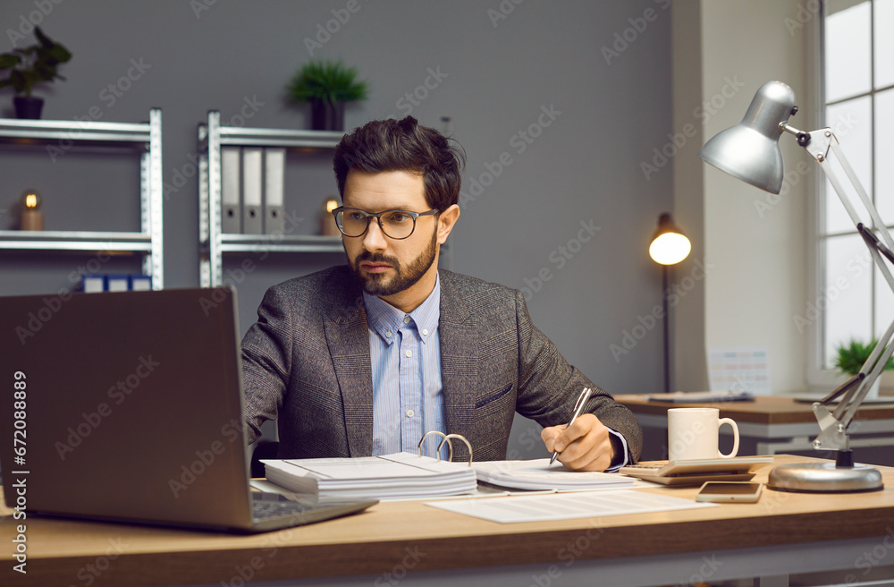 Focused businessman working on laptop and making notes on paper at workplace. Confident man in formal suit analyzing financial data, doing research while working in office Focused businessman working on laptop and making notes on paper at workplace. Confident man in formal suit analyzing financial data, doing research while working in office