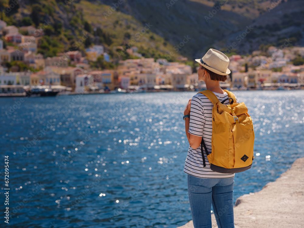 Nice asian Happy Female with backpack Enjoying her holidays on Symi Islands. View of port Symi or Simi, is tiny island of Dodecanese, Greece, calm atmosphere and fabulous architecture.
