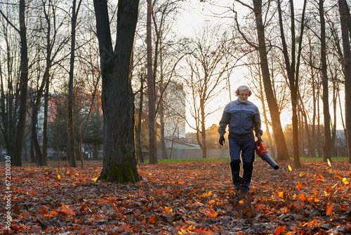 Serious senior man walking with handheld leaf blower to clean up park alley in October. Front view of focused bearded worker wearing uniform removing dry leaves in warm morning. Seasonal work concept.
