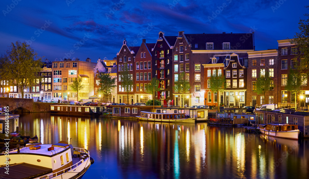 Evening town Amsterdam in Netherlands on bank river canal Amstel with shining window. Panorama landscape brown house over water reflection.