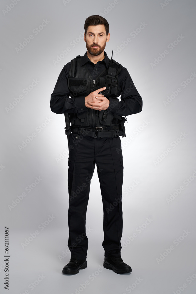 Attractive male cop with ammunition holding gun with both hands. Front view of bearded man in black body armor looking at camera, on gray studio background. Concept of work, police, rescue. Attractive male cop with ammunition holding gun with both hands. Front view of bearded man in black body armor looking at camera, on gray studio background. Concept of work, police, rescue.