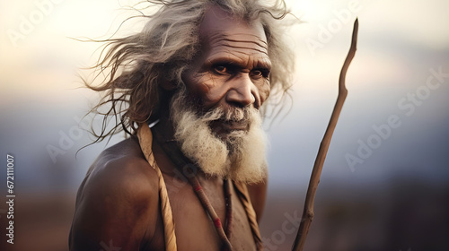 An Australian Aboriginal elder, with a rich tapestry of life experiences etched on his face, stands resolutely, clutching a staff against a tranquil outback backdrop. photo