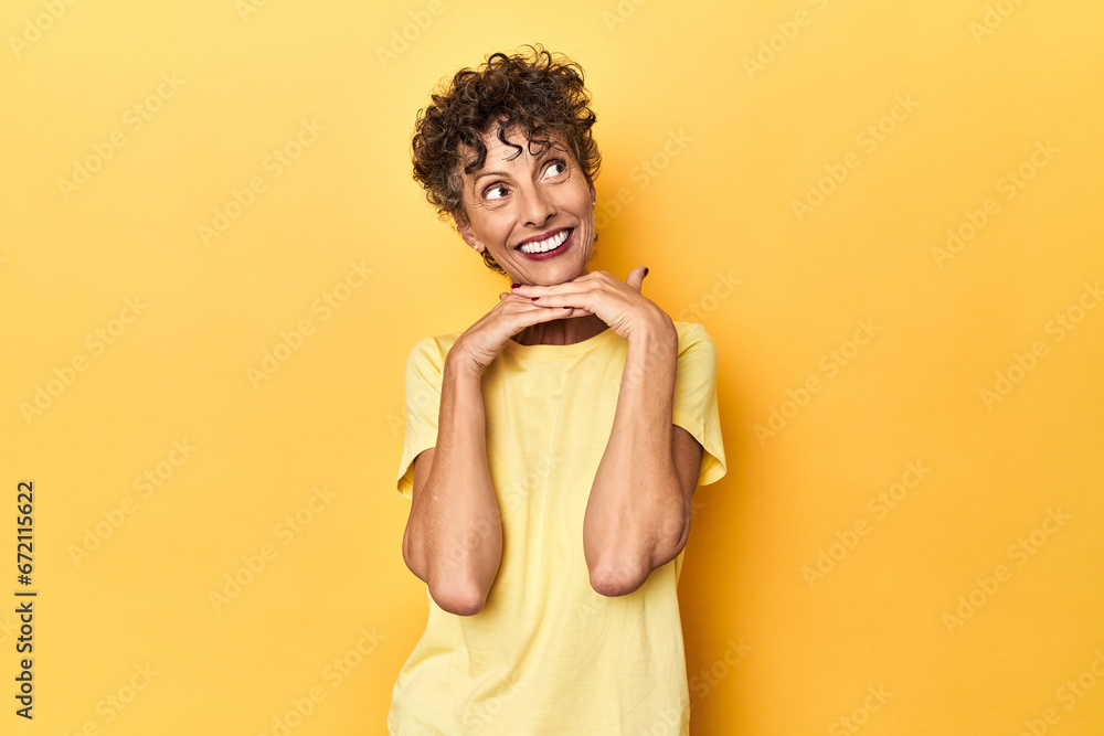 Mid-aged caucasian woman on vibrant yellow keeps hands under chin, is looking happily aside. Mid-aged caucasian woman on vibrant yellow keeps hands under chin, is looking happily aside.