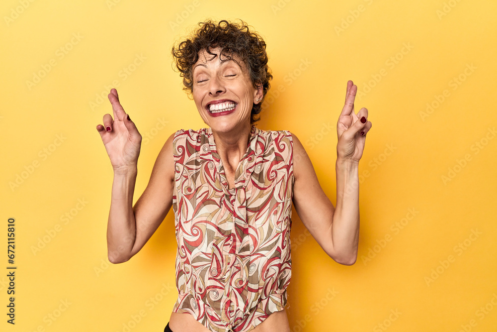 Mid-aged caucasian woman on vibrant yellow crossing fingers for having luck - obrazy, fototapety, plakaty 