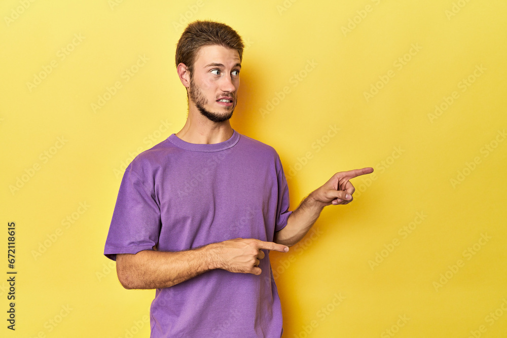 Young Caucasian man on a yellow studio background shocked pointing with index fingers to a copy space. Young Caucasian man on a yellow studio background shocked pointing with index fingers to a copy space.