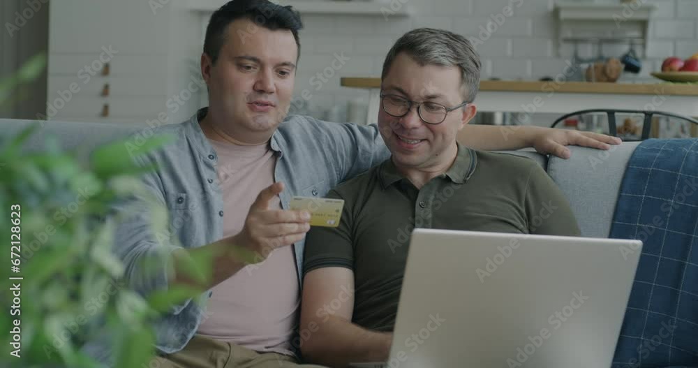 Cheerful same sex couple shopping online with laptop and credit card talking sitting on couch at home. Banking and e-commerce concept. Cheerful same sex couple shopping online with laptop and credit card talking sitting on couch at home. Banking and e-commerce concept.