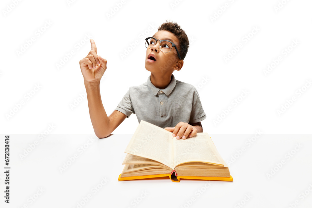 African-American boy, pupil sitting with opened book, create new idea during doing homework. Child study and learn for preschool. Online education. Concept of childhood, International Knowledge Day. African-American boy, pupil sitting with opened book, create new idea during doing homework. Child study and learn for preschool. Online education. Concept of childhood, International Knowledge Day.