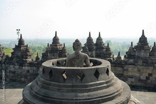 Obraz na płótnie (Selective focus) Stunning view of a Buddha Statue in the foreground and some bell shaped stupas in the background. Borobudur is a Mahayana Buddhist temple in Indonesia. w salonie