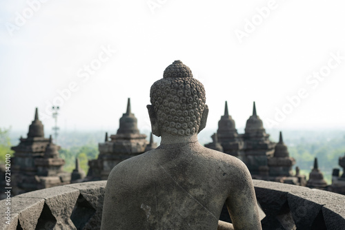Obraz na płótnie (Selective focus) Stunning view of a Buddha Statue in the foreground and some bell shaped stupas in the background. Borobudur is a Mahayana Buddhist temple in Indonesia. w salonie