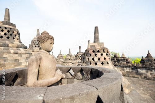 Obraz na płótnie (Selective focus) Stunning view of a Buddha Statue in the foreground and some bell shaped stupas in the background. Borobudur is a Mahayana Buddhist temple in Indonesia. w salonie