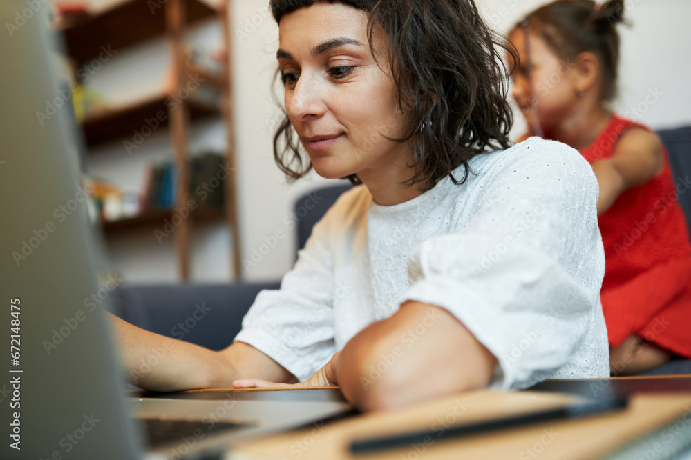 Fototapeta premium Portrait with selective focus on young mother freelancer, copywriter working on laptop or studying online from home while her little daughter watching tv on background sitting on couch